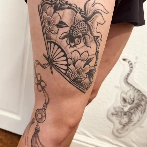 black and grey japanese folding fan with a goldfish, cherry blossoms and a hanging tassel, thigh piece, by hannah at shade studio peterborough
