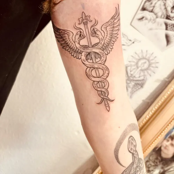fine line winged caduceus with two serpents coiled around a sword, back of the upper arm, by hannah at shade studio peterborough
